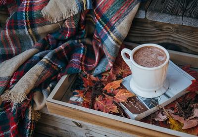 Hot chocolate resting on a book surrounded by autumn leaves and a tartan blanket